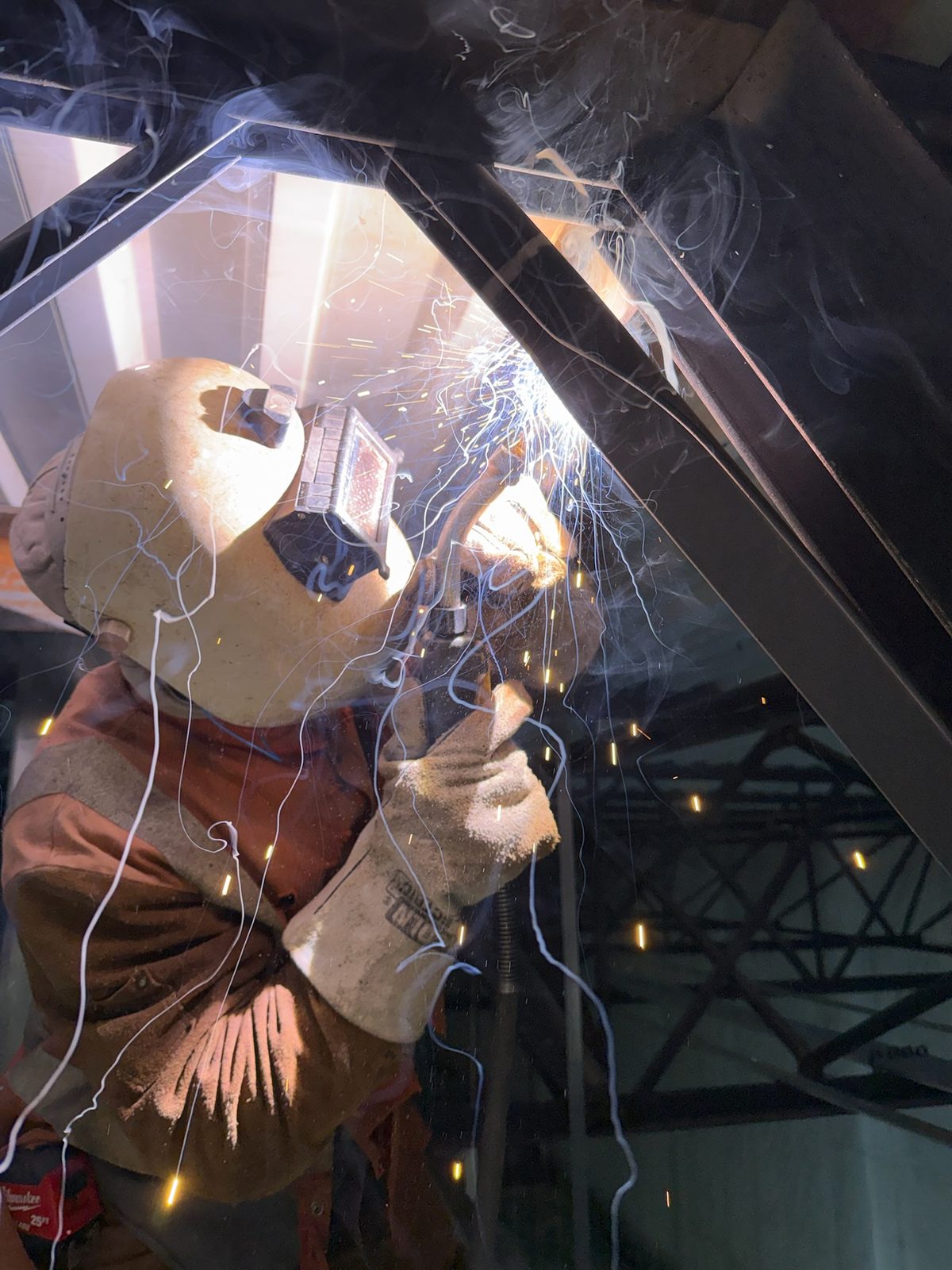 Welder in full leathers running an overhead weld on RTU reinforcement angles inside an occupied building